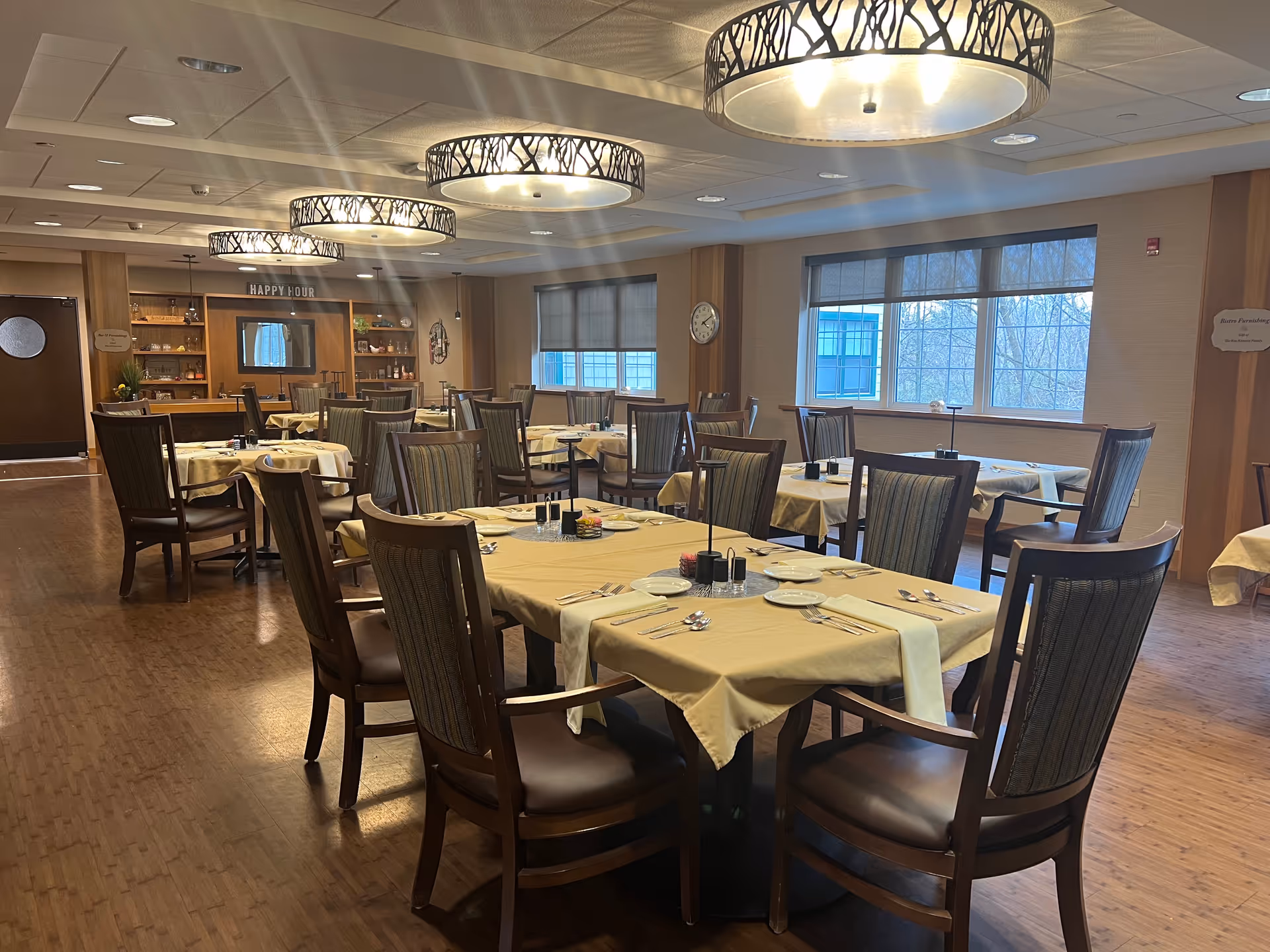 A dining room with multiple tables covered in beige tablecloths, each set with plates, silverware, and condiments. The room has large windows with blinds, wooden flooring, and decorative ceiling lights. In the background, there is a bar area with a 'HAPPY HOUR' sign above it.