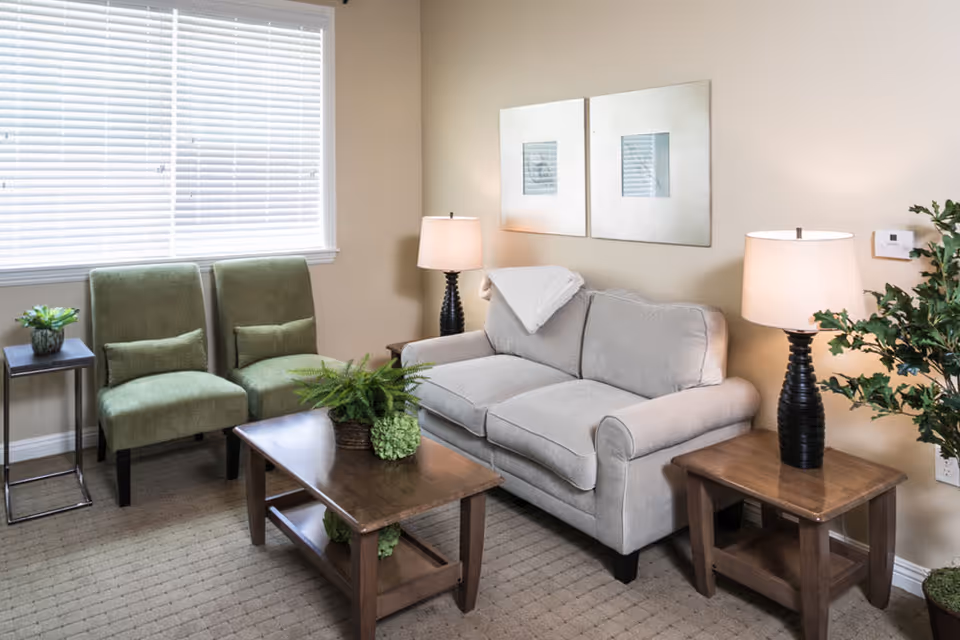 Bright sitting area with a beige loveseat, two green chairs, wooden coffee and side tables, lamps, and plants.