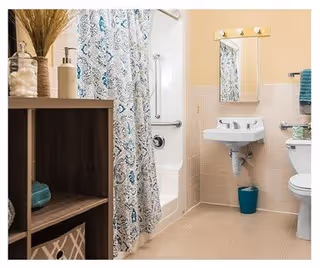 A clean and well-lit bathroom featuring a white sink with a mirror above it, a toilet, a bathtub with a patterned shower curtain, and a wooden shelving unit holding towels and decorative items.