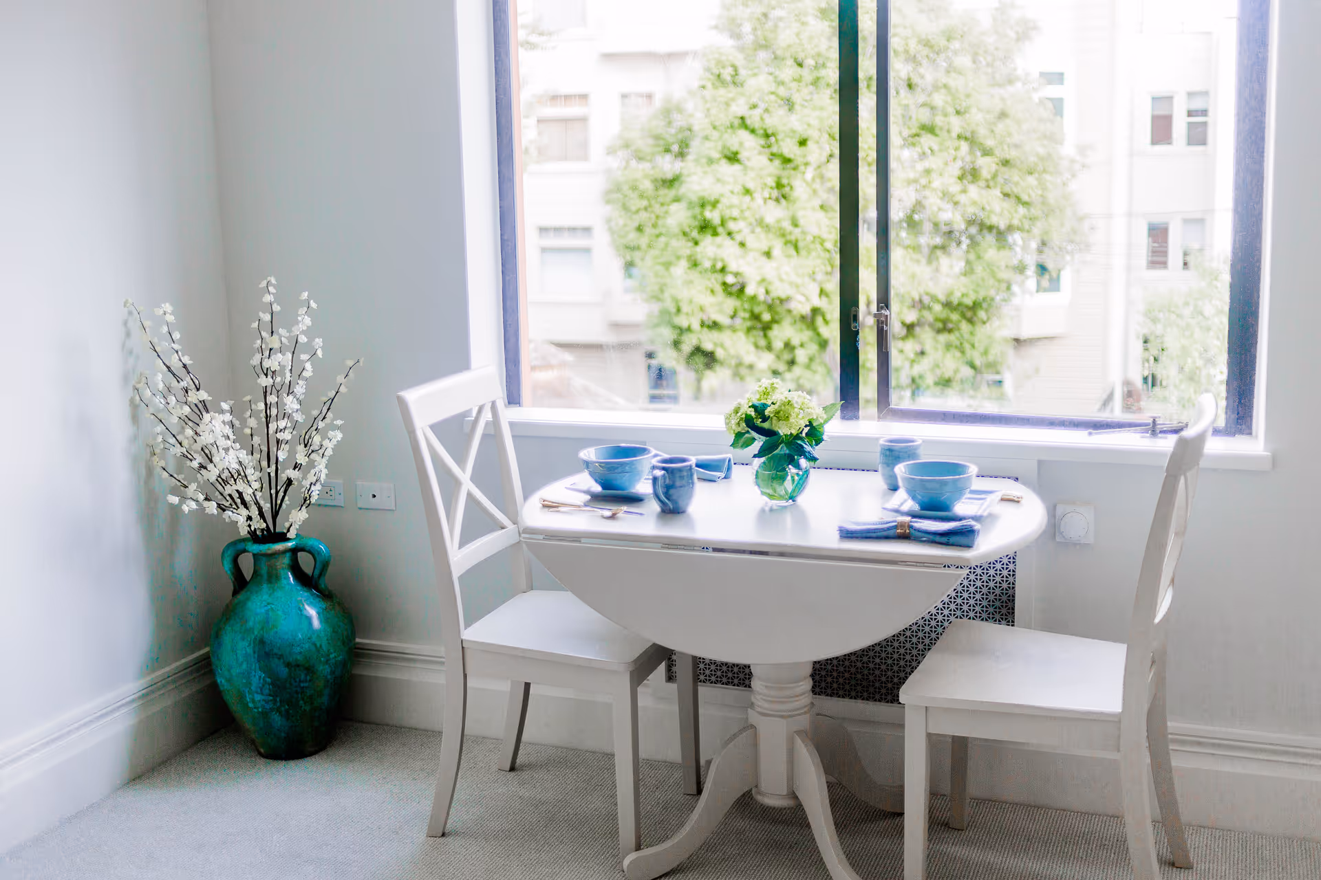 Bright dining nook with a small white round table set for two by a large window and a turquoise floor vase with white branches.