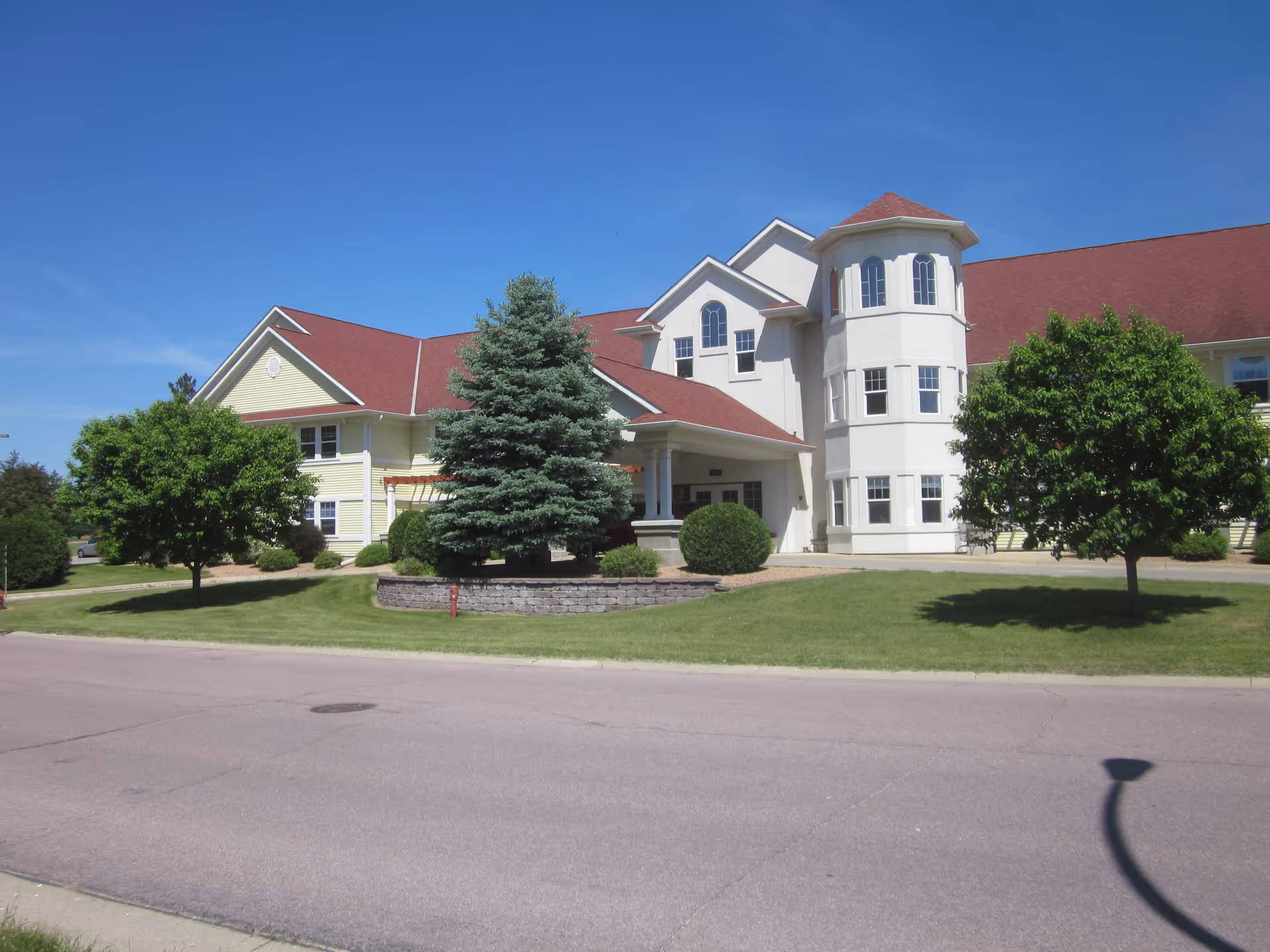 Front exterior of a large two-story senior living building with a red roof, turret, trees and a manicured lawn under a clear blue sky.