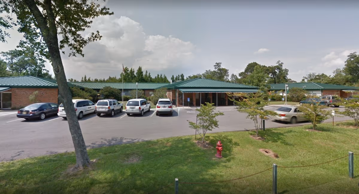 One-story brick nursing center with green metal roofs, a parking lot of cars, trees, and a fire hydrant in the foreground.
