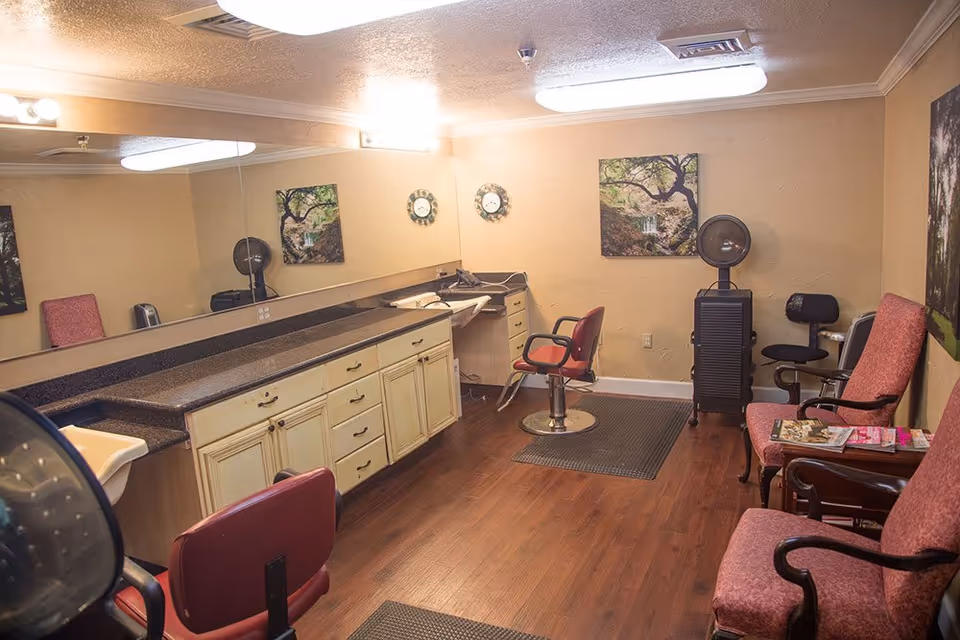 Interior of a hair salon room with wooden flooring, a large mirror covering one wall, a countertop with drawers, salon chairs, a hair dryer, and a few framed pictures on the walls. There are also two upholstered chairs with magazines on a small table between them.