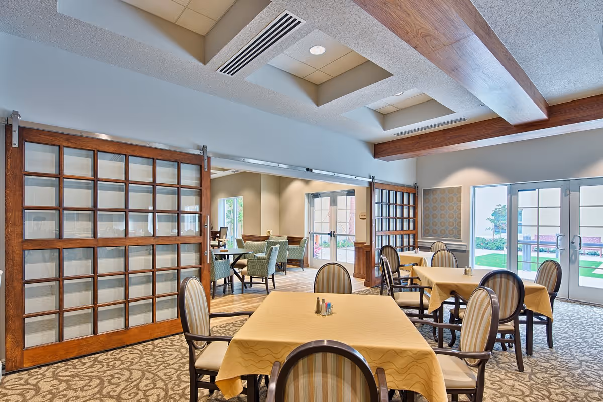 A bright dining room with several tables covered in yellow tablecloths and surrounded by striped upholstered chairs. The room features large wooden sliding doors with glass panels, patterned carpet, and ceiling with recessed lighting. Double glass doors lead outside to a green lawn area.