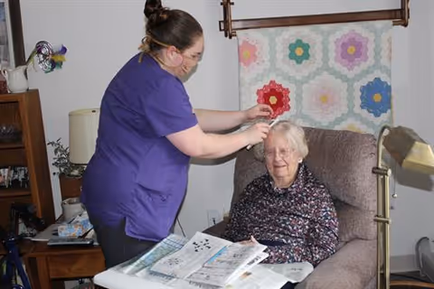A caregiver wearing a purple uniform and a face mask is styling the hair of an elderly woman seated in a comfortable armchair in a cozy living room. The elderly woman is smiling and holding a newspaper. The room features a quilt hanging on the wall, a table lamp, and various household items on a side table.