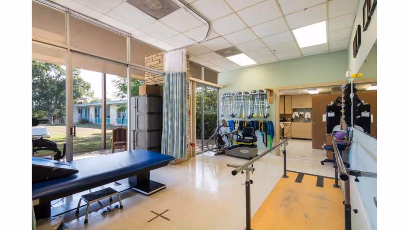 Physical therapy room with parallel bars, exercise equipment, a treatment table, and large windows showing an outdoor view with trees and a building.