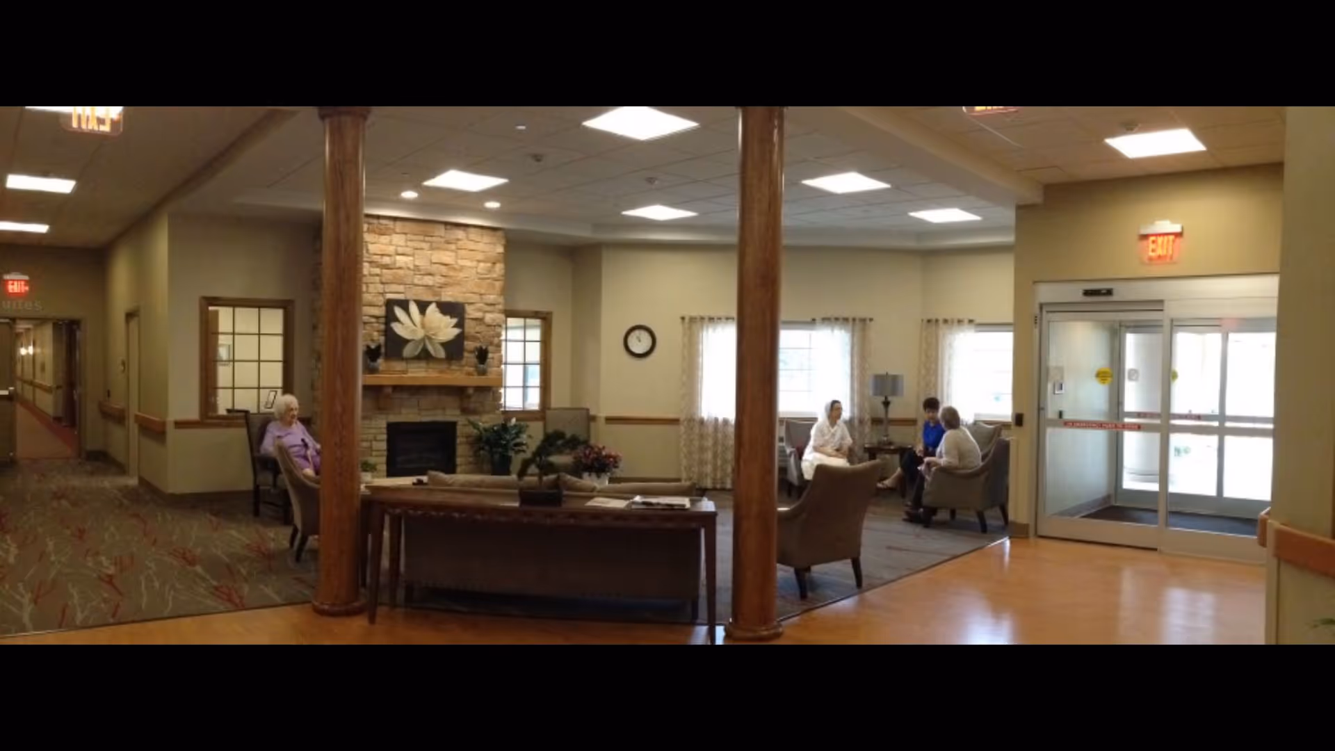 A cozy senior living facility common area with a stone fireplace, wooden columns, and comfortable seating. Several elderly individuals are seated and engaged in conversation. The room has large windows with curtains, a clock on the wall, and an exit door leading outside.