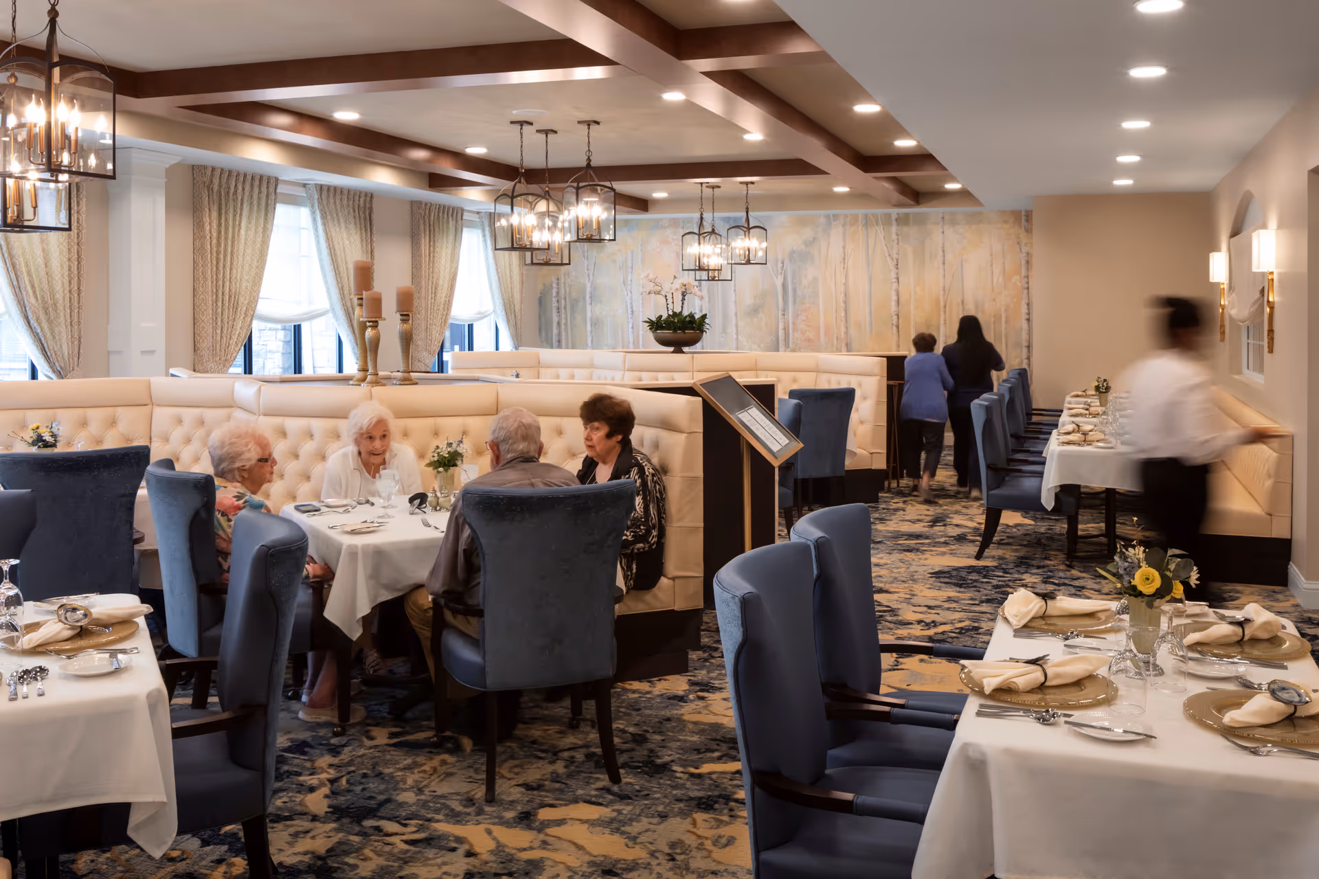 Elegant dining room with elderly residents seated at tables and tufted booths under chandeliers.
