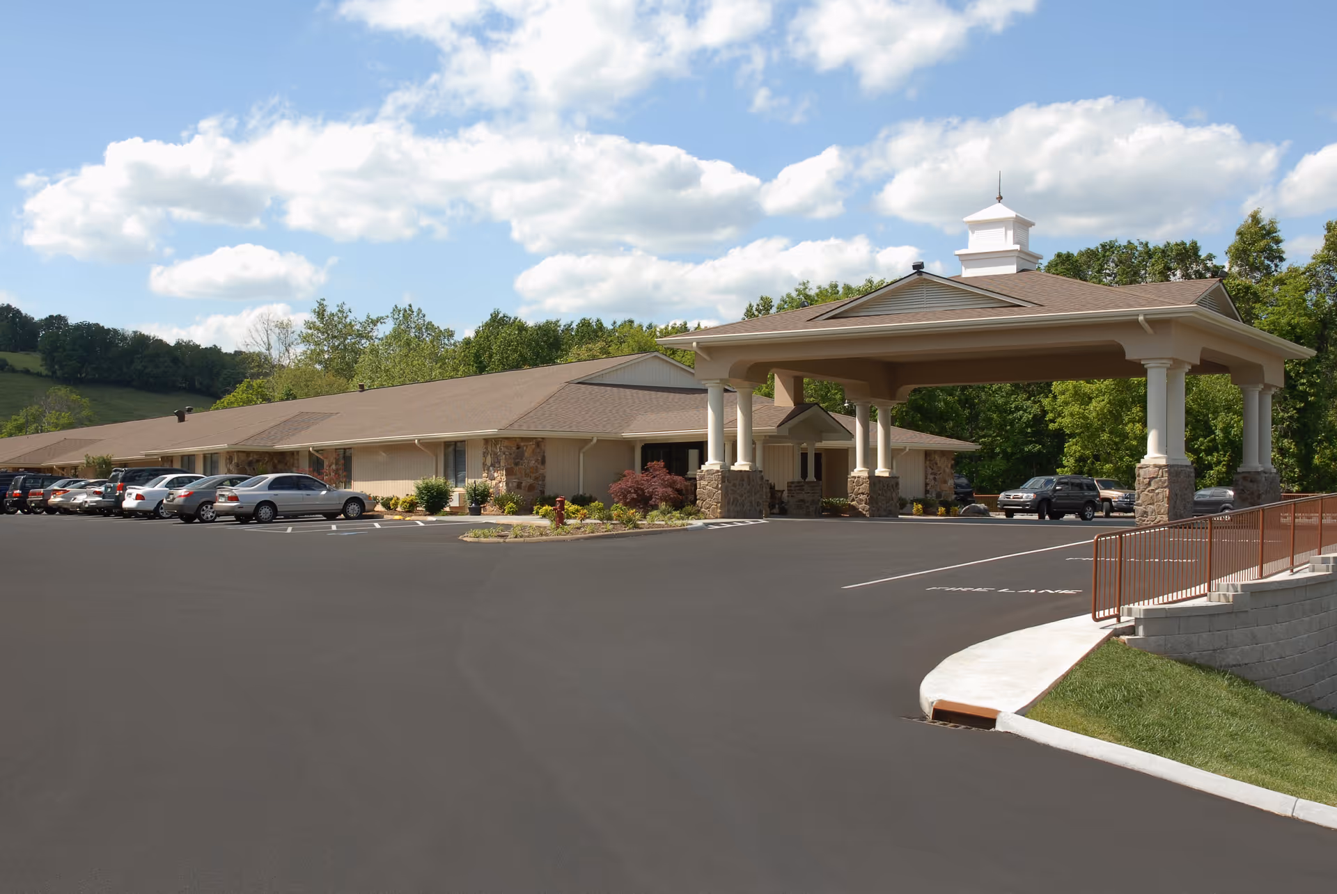 Exterior view of a single-story senior living facility building with a covered entrance supported by columns. Several cars are parked in the parking lot in front of the building. The sky is partly cloudy and there are trees and greenery surrounding the facility.