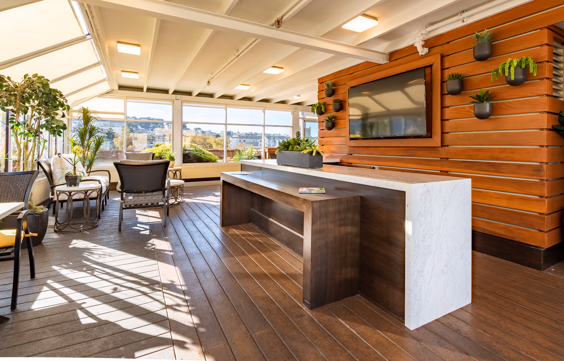 Bright indoor lounge area with large windows allowing natural light to flood the space. The room features wooden flooring, a long wooden table with a white marble side, several chairs and small tables with potted plants, and a wooden wall with a mounted flat-screen TV and small wall-mounted planters.