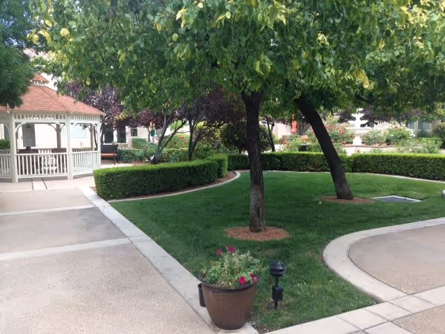 A well-maintained outdoor garden area with green grass, trimmed hedges, and several trees. There is a white gazebo on the left side and a curved walkway surrounding the garden. A potted plant with flowers is placed near the walkway.