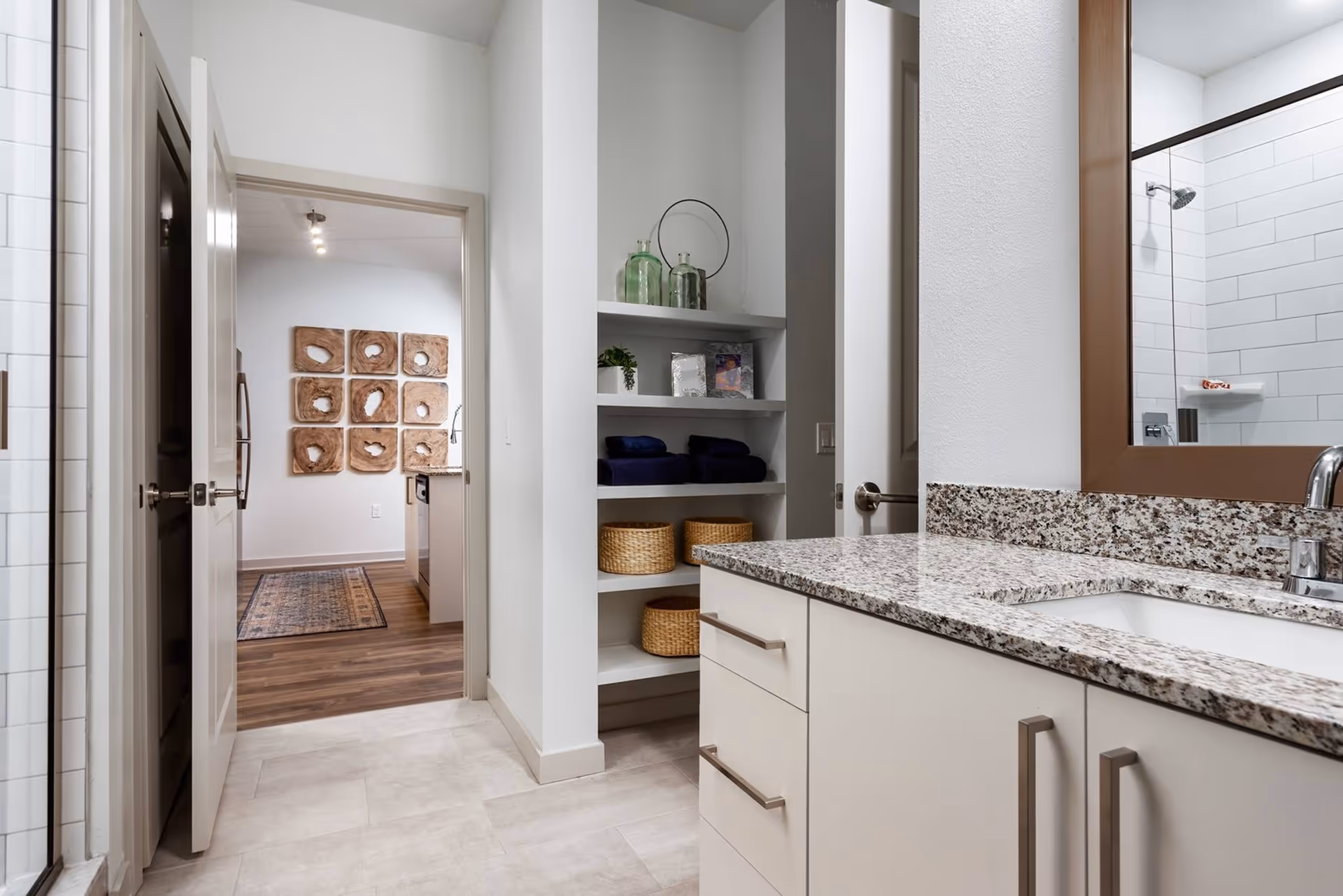 A bright bathroom with a granite-topped vanity and sink, mirror and glass shower to the right, built-in shelving with baskets, and an open door leading to a hallway with wall art.
