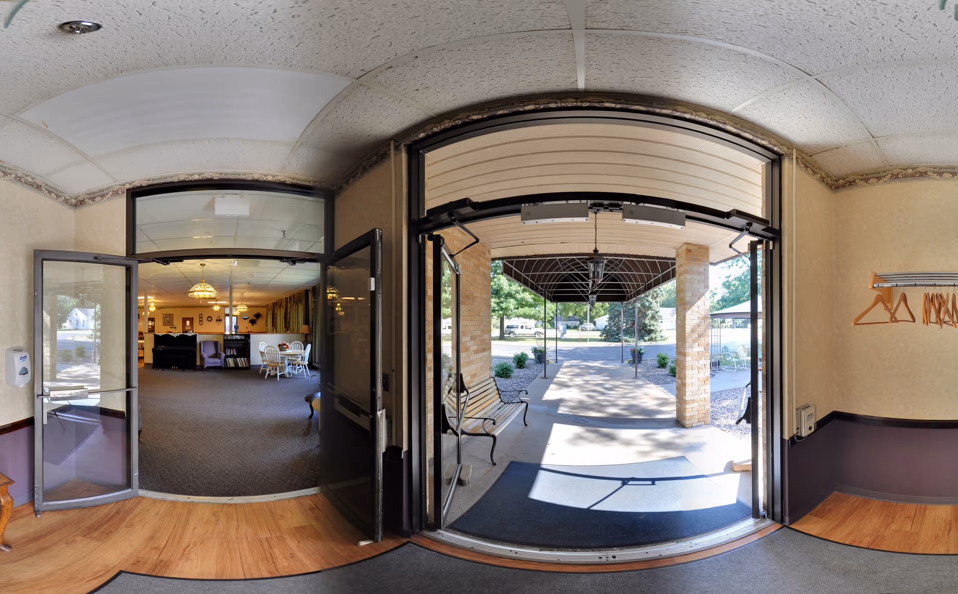 View from an entrance area looking out to a covered outdoor walkway with benches and greenery. Inside, there is a room with carpeted flooring, tables, chairs, and a chandelier, suggesting a common or lounge area.