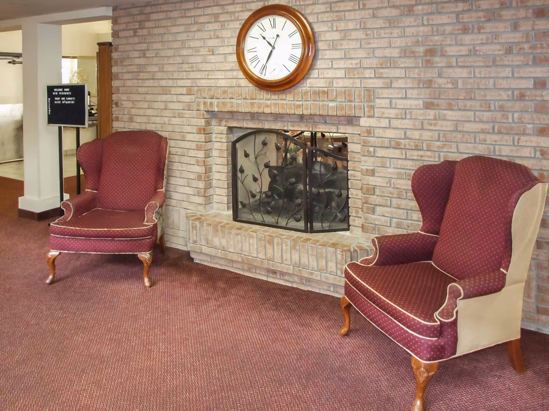 Two maroon upholstered wingback chairs with wooden legs positioned on either side of a brick fireplace with a decorative metal screen. Above the fireplace is a round wall clock with Roman numerals. The room has a maroon carpet and a small signboard in the background near a doorway.