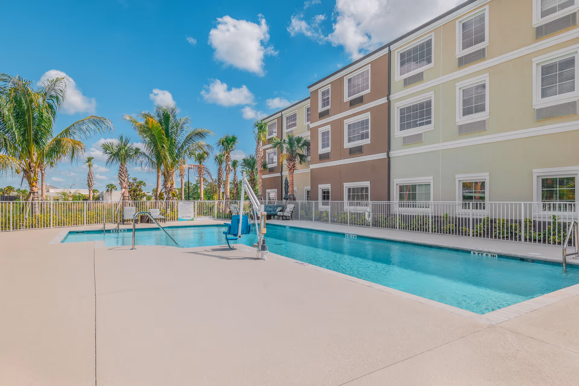 Outdoor swimming pool area at a senior living facility with clear blue water, surrounded by a safety fence. Palm trees and a multi-story building with beige and light green walls and multiple windows are visible in the background under a blue sky with some clouds.
