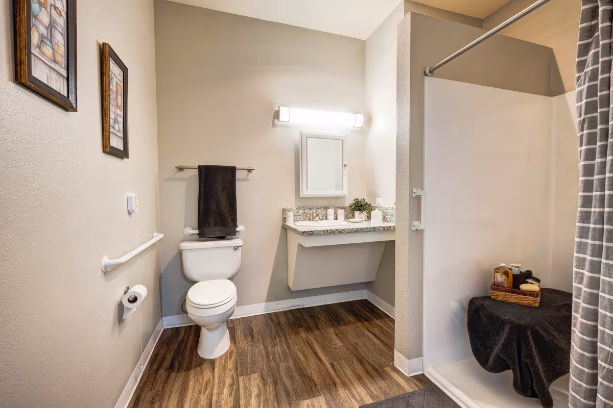 A clean and modern bathroom with a toilet, a wall-mounted towel rack holding a black towel, a floating vanity with a granite countertop, a small potted plant, and a mirrored medicine cabinet above. There is a walk-in shower with a gray and white striped curtain and a small bench inside holding a basket with toiletries. The floor has wood-style vinyl planks and the walls are painted light beige with two framed pictures hanging.