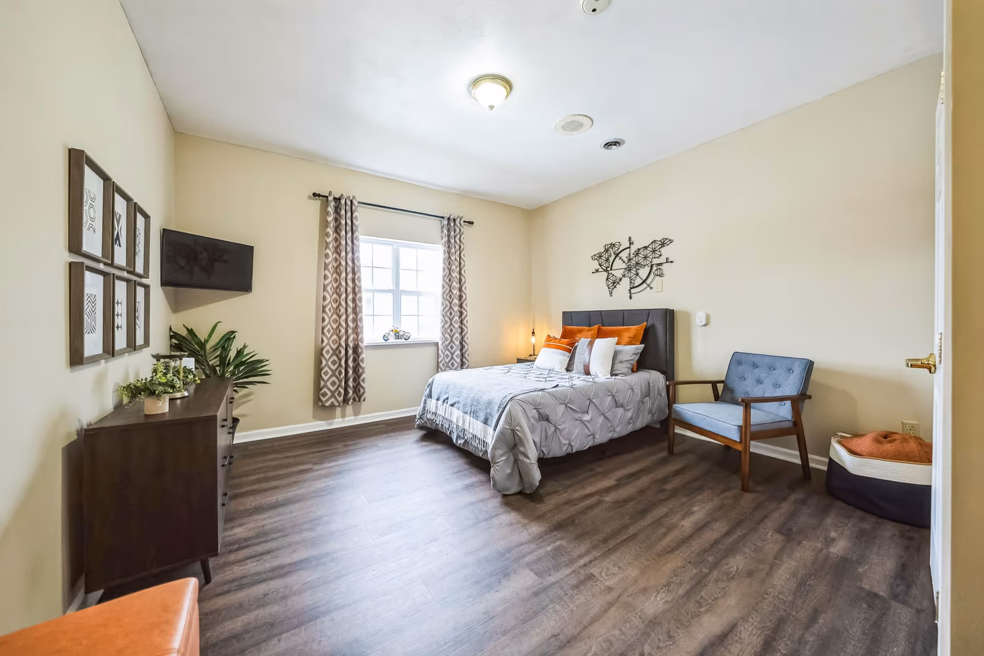 A bright bedroom with a large window covered by patterned curtains. The room features a bed with gray bedding and orange accent pillows, a blue armchair, a dark wooden dresser with plants and decor, and framed artwork on the wall. The floor is wood, and the walls are painted light beige.