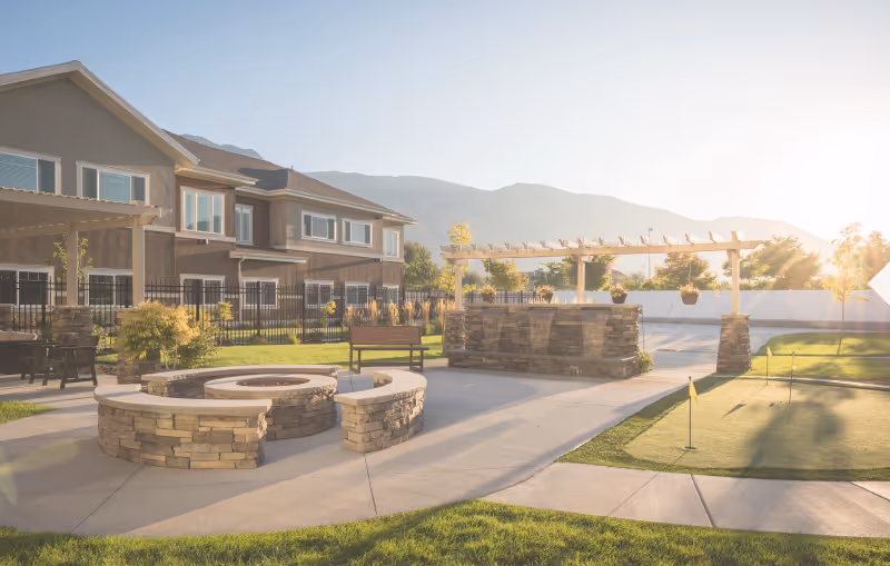 Outdoor patio area at Ashford Assisted Living & Memory Care featuring a stone fire pit with curved seating, a pergola with hanging plants, benches, and a putting green, with a two-story building and mountains in the background under a bright sky.