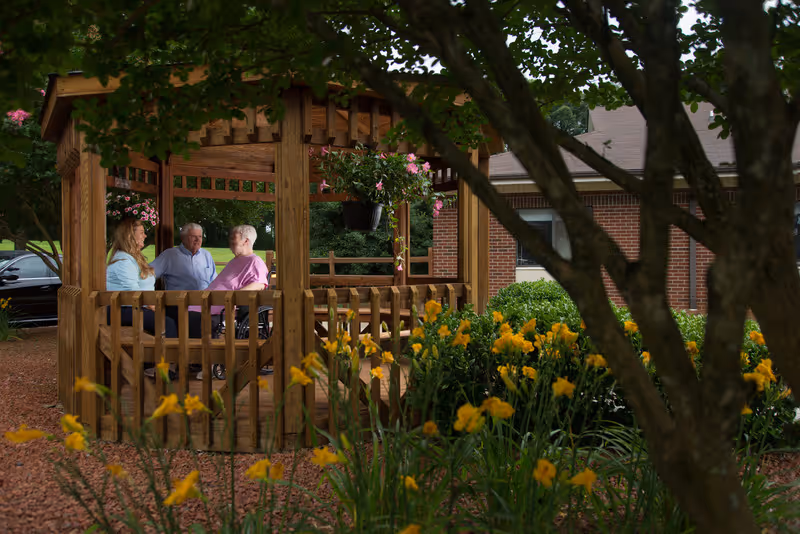 Three people sitting and talking inside a wooden gazebo surrounded by greenery and yellow flowers, with a brick building visible in the background.