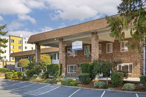 Exterior view of Brookdale Federal Way senior living facility showing a brick building with a covered entrance, American and Washington state flags, well-maintained shrubs, and a parking area in front under a partly cloudy sky.