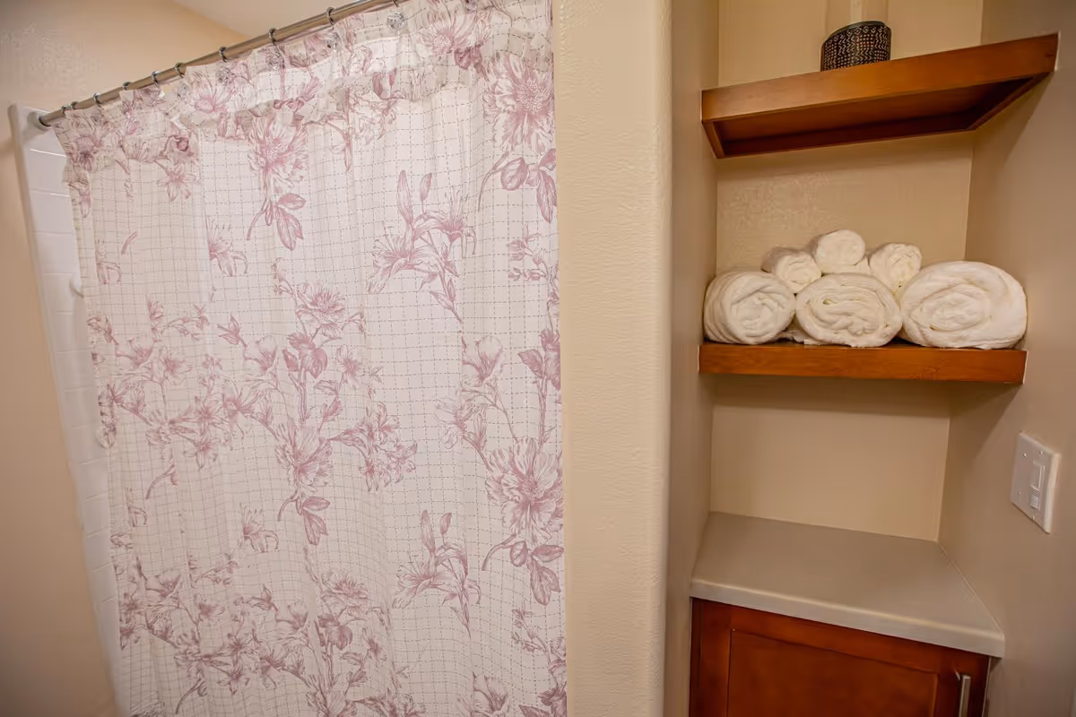 Bathroom corner with a floral shower curtain and wooden shelves holding rolled white towels.