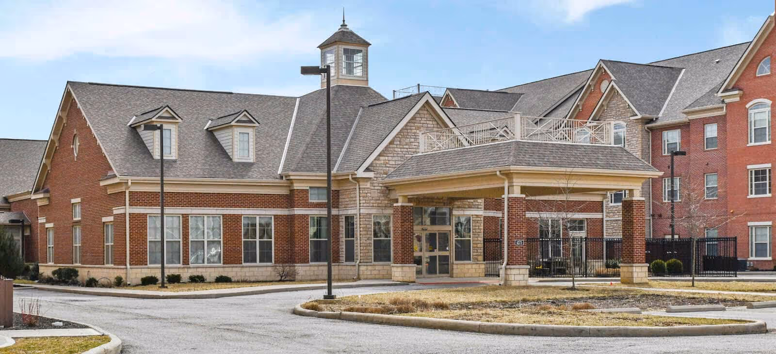 Exterior view of a senior living facility named Avondale, featuring a brick building with multiple windows, a covered entrance with columns, and a cupola on the roof. The surrounding area includes a driveway and some landscaping with grass and small shrubs.