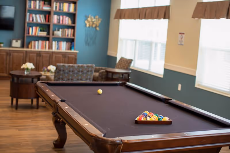 A cozy indoor recreational room featuring a pool table with a full rack of colorful billiard balls and a cue ball. In the background, there is a bookshelf filled with books, a couple of armchairs, and a small round table with flower arrangements. The room has wooden flooring and large windows with brown valances allowing natural light to brighten the space.