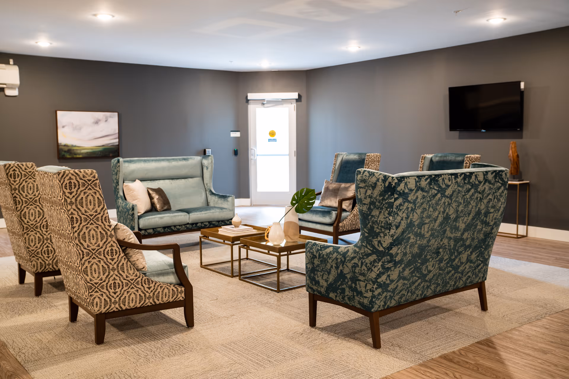 A modern and cozy living room area with patterned and solid upholstered armchairs and loveseats arranged around two small square coffee tables. The room has gray walls, a light carpet, a wall-mounted flat screen TV, a piece of artwork on the wall, and a door with a window letting in natural light.
