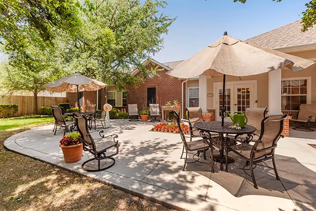 Outdoor patio area at Brookdale Westover Hills with metal tables and chairs under large beige umbrellas, surrounded by trees and a brick building in the background.