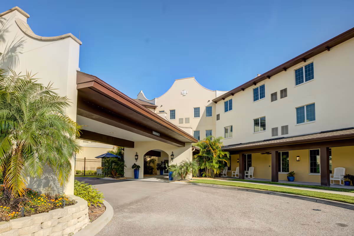 Exterior view of Arbor Terrace Citrus Park senior living facility showing a beige multi-story building with multiple windows, a covered entrance with a brown roof, palm trees, and outdoor seating with white chairs under a covered walkway.