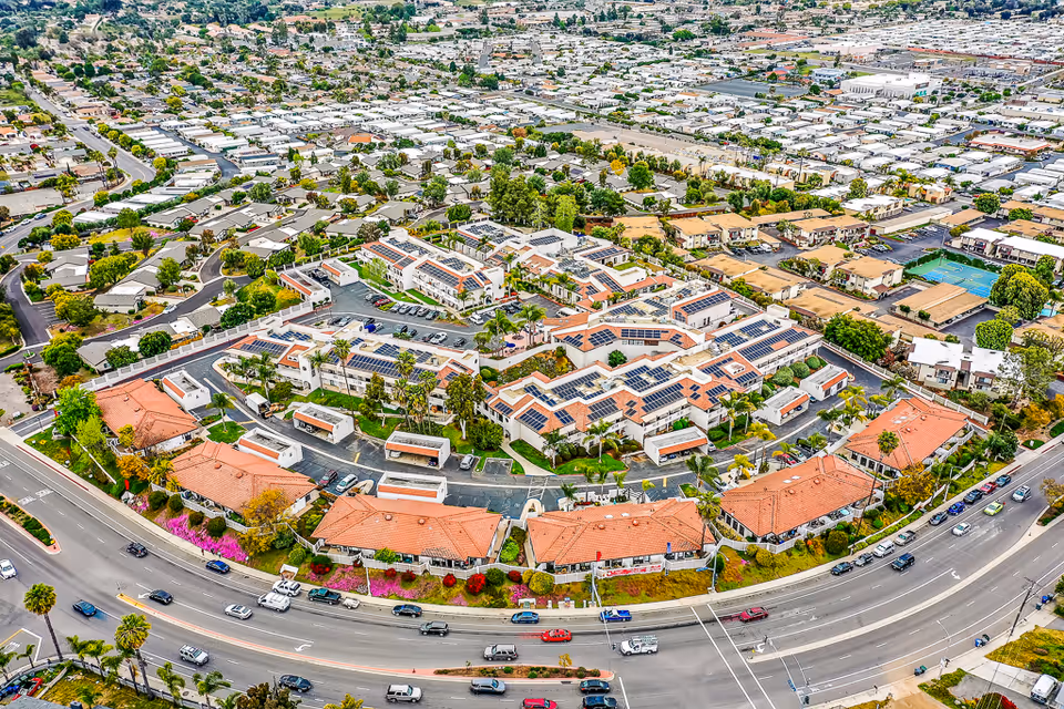 Aerial view of Rancho Vista Senior Living campus with multiple terracotta-roofed buildings, solar panels, landscaped grounds, parking areas, and surrounding neighborhood streets.