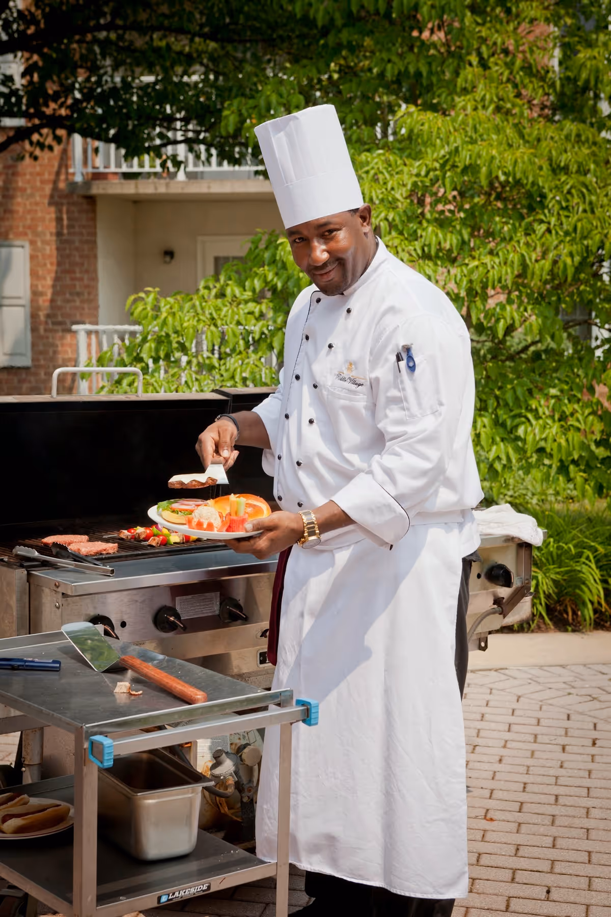 A chef wearing a white uniform and tall chef hat is grilling food outdoors. He is holding a plate with grilled vegetables and meat, and is using a spatula to place food on the plate. Behind him is a barbecue grill with food cooking, and green trees and a building are visible in the background.