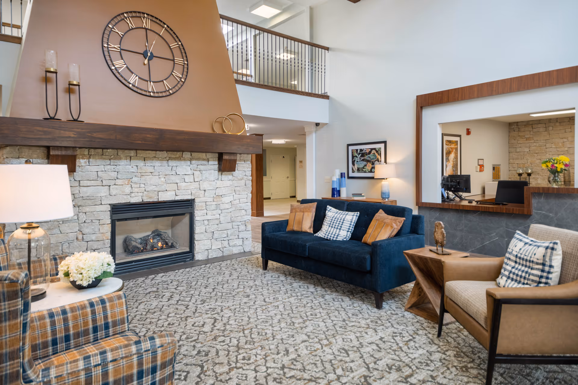 A cozy living room area in a senior living facility featuring a stone fireplace with a large clock above it, a blue sofa with pillows, a plaid armchair, a beige armchair with a plaid pillow, a patterned carpet, and a reception desk visible through a large window opening in the wall.