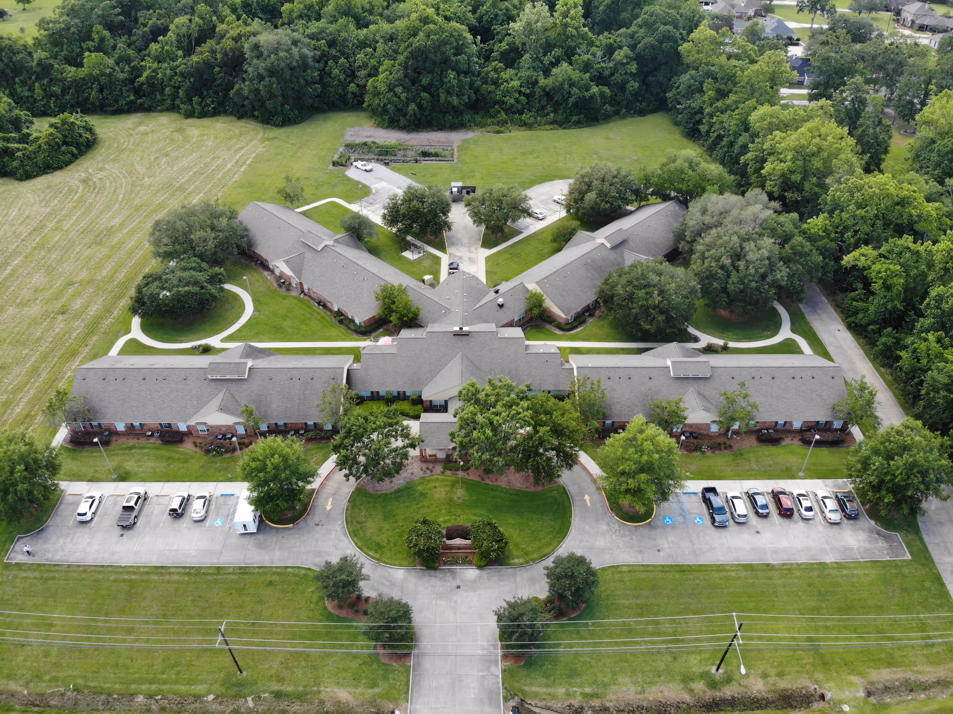 Aerial view of Azalea Estates of Gonzales, showing a large single-story building complex surrounded by green lawns, trees, and parking lots with several cars. The building is shaped with multiple wings extending from a central area, and there are pathways and driveways around the property.
