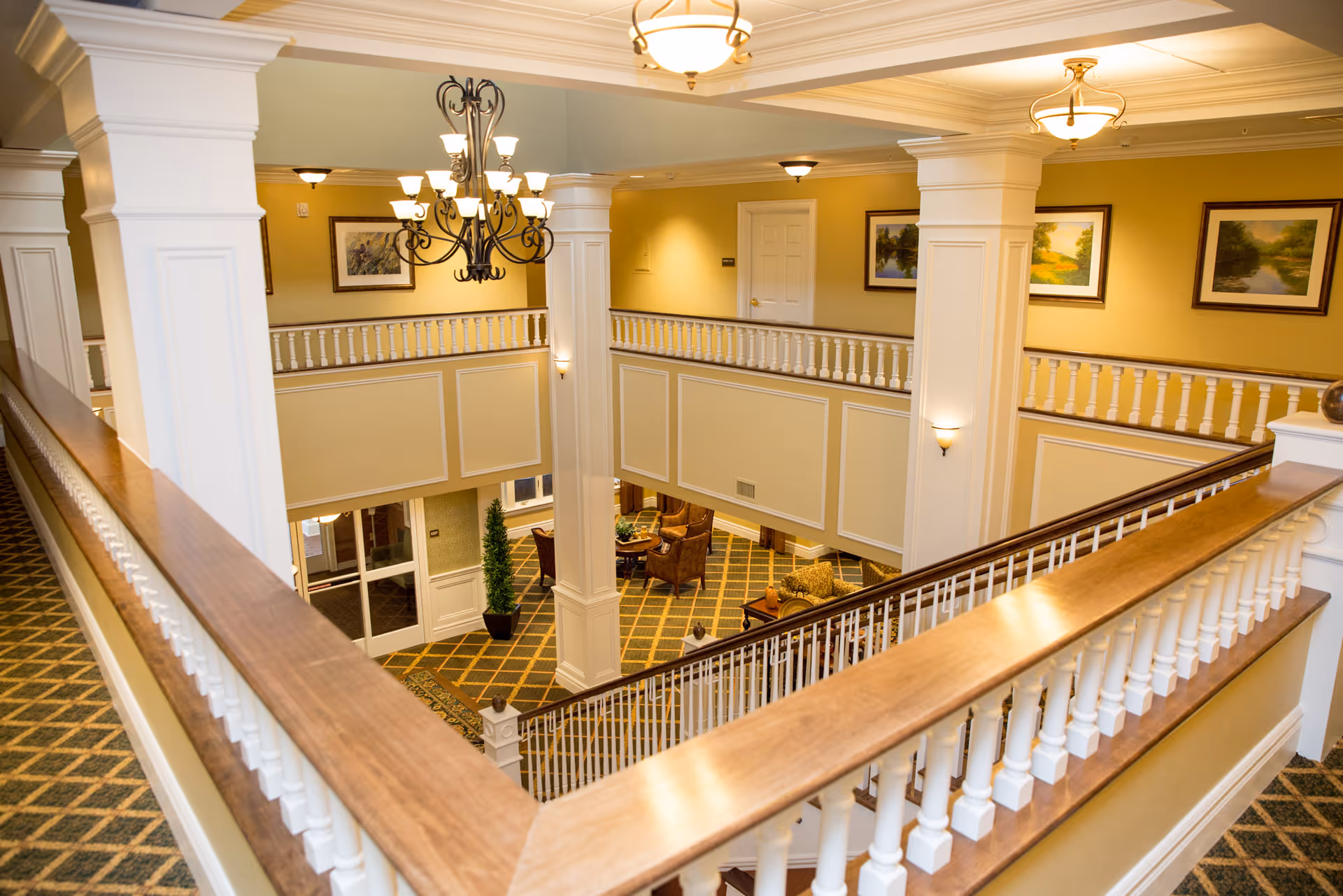 Interior view of a two-story common area in a retirement community with white columns, wooden railings, chandeliers, framed landscape paintings on yellow walls, and a seating area with chairs and a table on the lower level.
