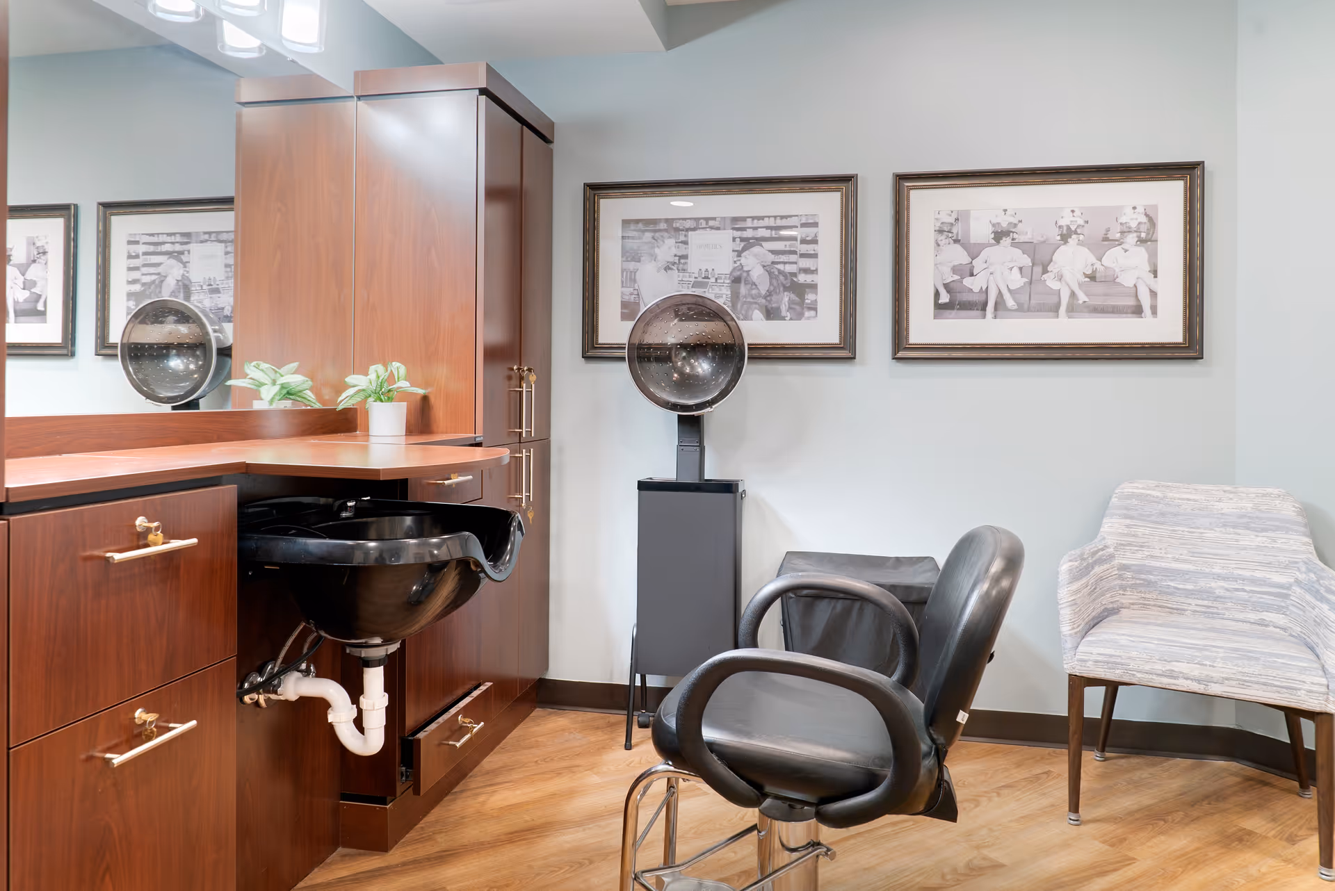 A salon-style room with a styling chair and black wash basin set against wood cabinetry, a hood dryer, framed pictures, and extra seating.
