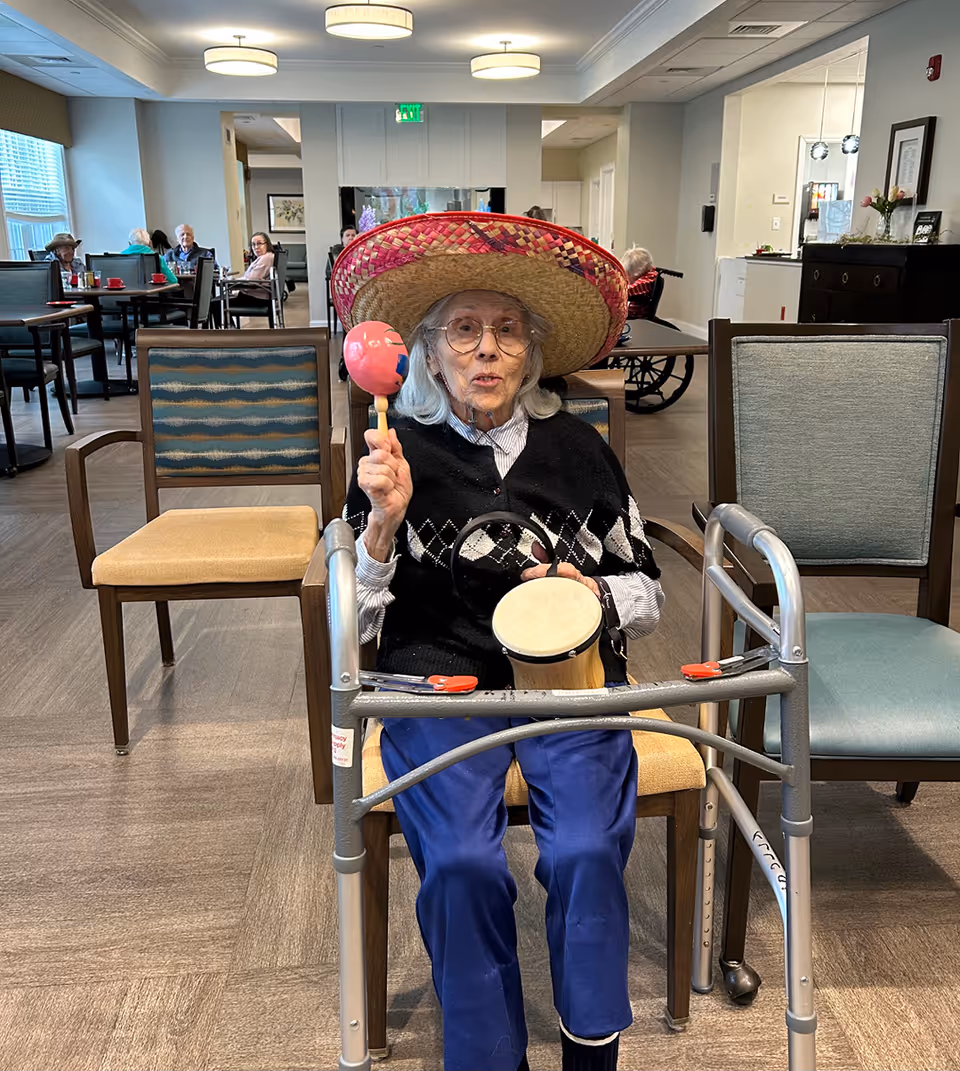 An elderly woman wearing a large colorful sombrero sits in a chair in a common room of a senior living facility. She holds a maraca in one hand and a small drum in the other. A walker is positioned in front of her. In the background, other elderly residents are seated at tables, and the room is well-lit with ceiling lights and large windows.