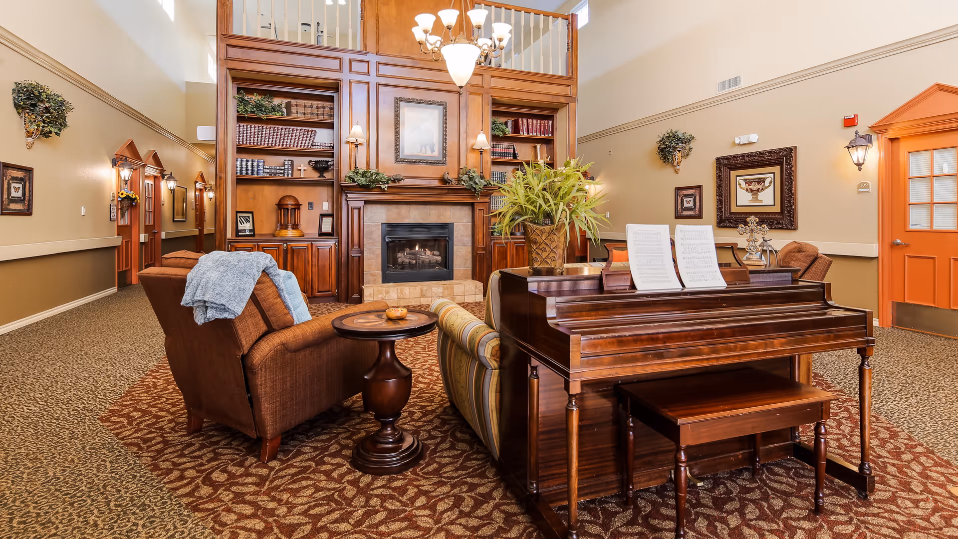 A cozy living room area in a senior living facility featuring a wooden piano with sheet music, a striped sofa, a brown armchair with a blue throw blanket, a round wooden side table, and a fireplace with built-in wooden shelves filled with books and decorative items. The room has warm beige walls, framed artwork, wall sconces, and a patterned carpet. There is an orange door on the right side and a hallway with additional doors and wall decorations on the left.