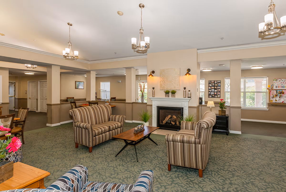 A senior living facility common area with striped upholstered chairs and a loveseat arranged around a wooden coffee table in front of a white fireplace. The room has beige walls, green patterned carpet, and multiple ceiling light fixtures. There are plants on the mantel and side tables, and windows letting in natural light.