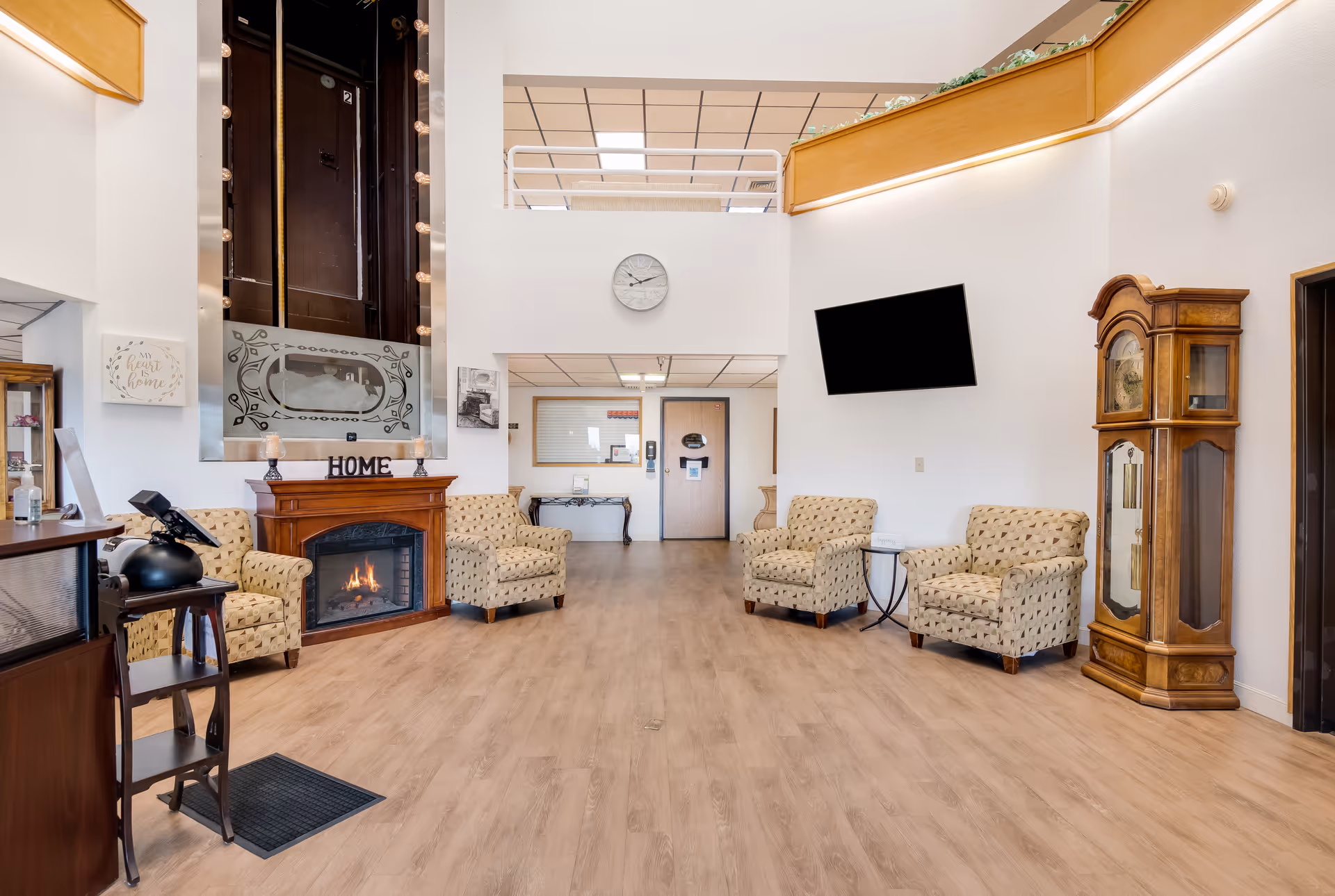 A bright and spacious senior living facility common area with light wood flooring, four patterned armchairs arranged around a wooden electric fireplace with a 'HOME' sign on the mantel. Above the fireplace is a vintage elevator door with decorative glass. A large wall clock is mounted on a white wall, and a flat-screen TV is angled on another wall. There is a tall wooden grandfather clock on the right side and a reception desk partially visible on the left. The ceiling has a wooden trim with recessed lighting and an upper balcony railing.