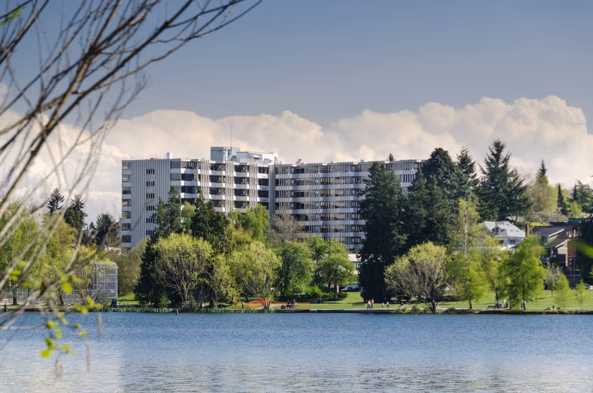 A large multi-story senior housing building named Village Cove is visible across a body of water, surrounded by lush green trees and a park area with people walking along the shoreline under a partly cloudy sky.