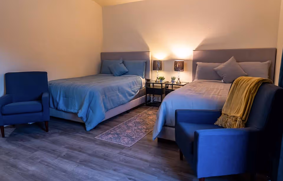 A softly lit bedroom with two twin beds, a shared nightstand with lamps, and two blue armchairs.