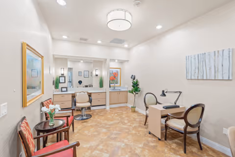 A well-lit interior room with beige walls and tiled floor, featuring two red cushioned chairs with a small round table holding a vase of flowers, a desk with two chairs and a desk lamp, and a vanity area with two mirrors, chairs, and framed artwork on the walls.