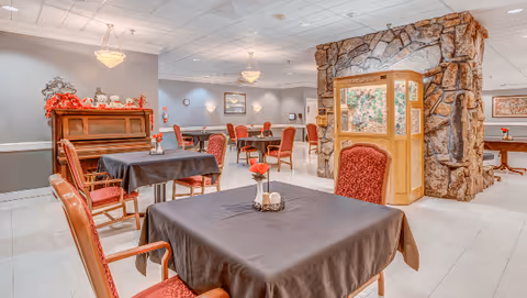 Spacious dining room with tables covered in dark tablecloths, red upholstered chairs, a piano, and a decorative stone partition.