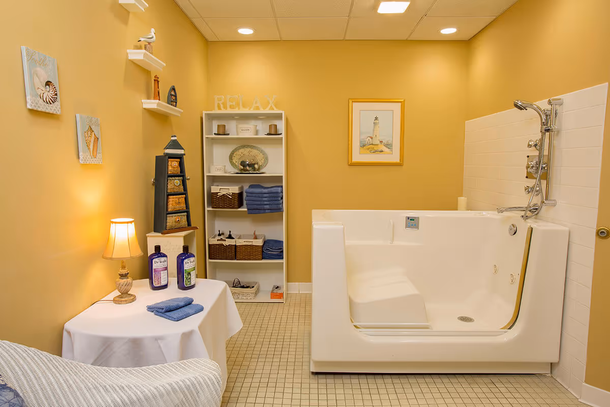 A cozy bathroom with yellow walls featuring a walk-in bathtub with a built-in seat and handheld showerhead. There is a white shelving unit stocked with towels and decorative items, a small table with a lamp and two bottles of bath products, and wall art including seashell pictures and a framed lighthouse painting. The floor is tiled with small square tiles.