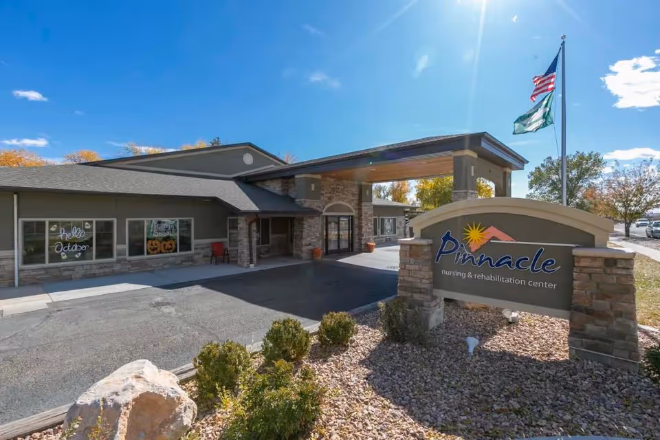 Exterior view of Pinnacle Nursing and Rehabilitation Center building with a covered entrance, stone pillars, and a sign displaying the facility's name. There are two flags on a flagpole and some landscaping with bushes and rocks in the foreground under a clear blue sky.