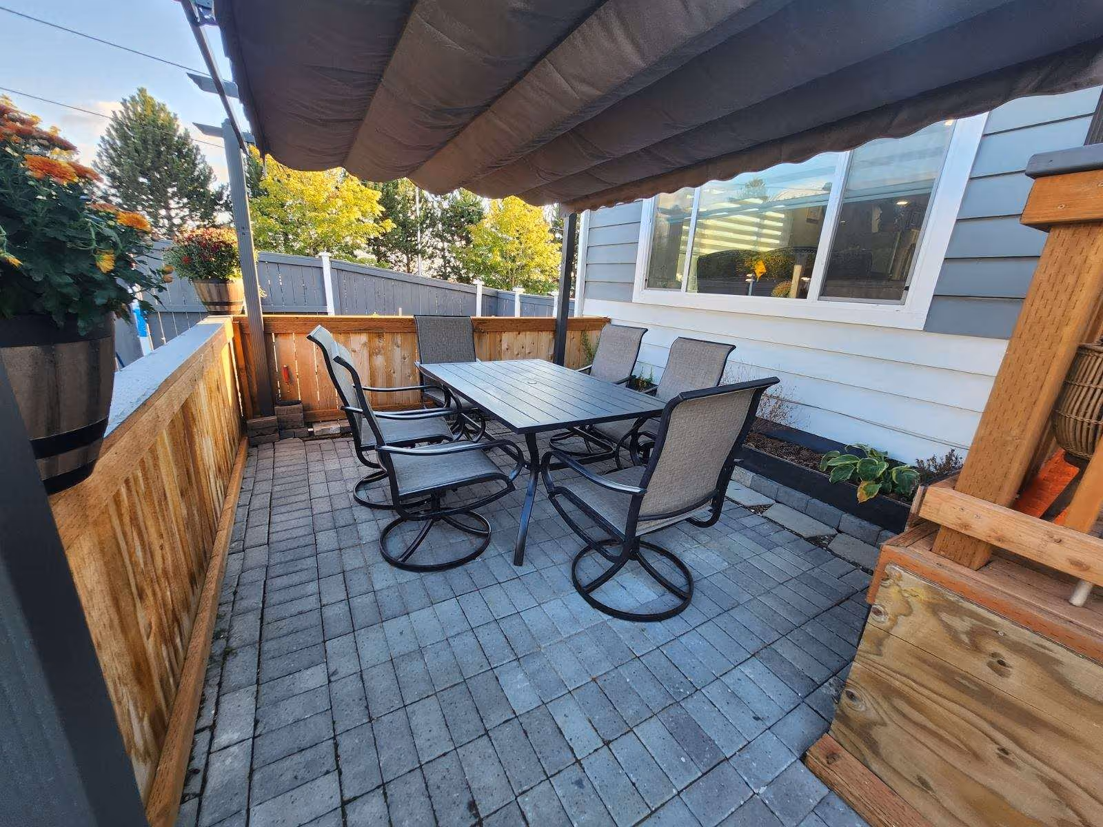 Outdoor patio area with a rectangular table and six metal chairs with mesh backs and seats. The patio is paved with square tiles and has a wooden fence on one side and a house wall with a window on the other. There is a retractable fabric canopy overhead providing shade. Potted plants and greenery are visible around the patio.