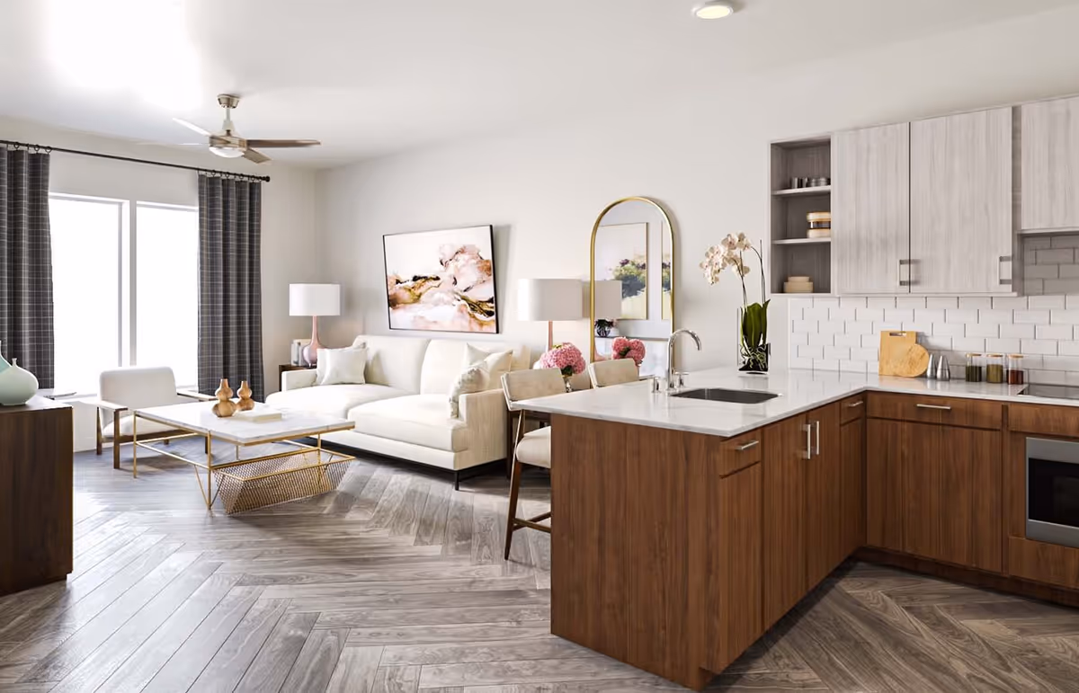 A modern open-concept living room and kitchen area with light wood flooring arranged in a herringbone pattern. The living room features a white sectional sofa, a white armchair, a marble coffee table with gold accents, a floor lamp, and a large abstract painting on the wall. The kitchen has wooden lower cabinets, light upper cabinets, a white subway tile backsplash, a white countertop with a sink, and two bar stools. There is a tall arched mirror and a vase with flowers on the kitchen counter.