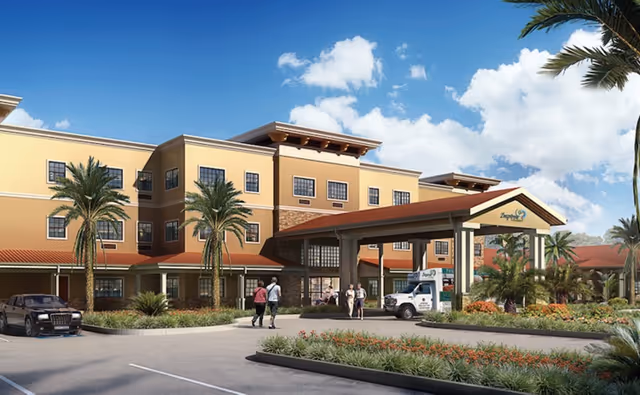 Exterior view of a senior living facility with a three-story building featuring yellow and beige walls, red-tiled roof, and palm trees in the landscaped front area. Several people are walking near the entrance, and a vehicle is parked under a covered drop-off area.