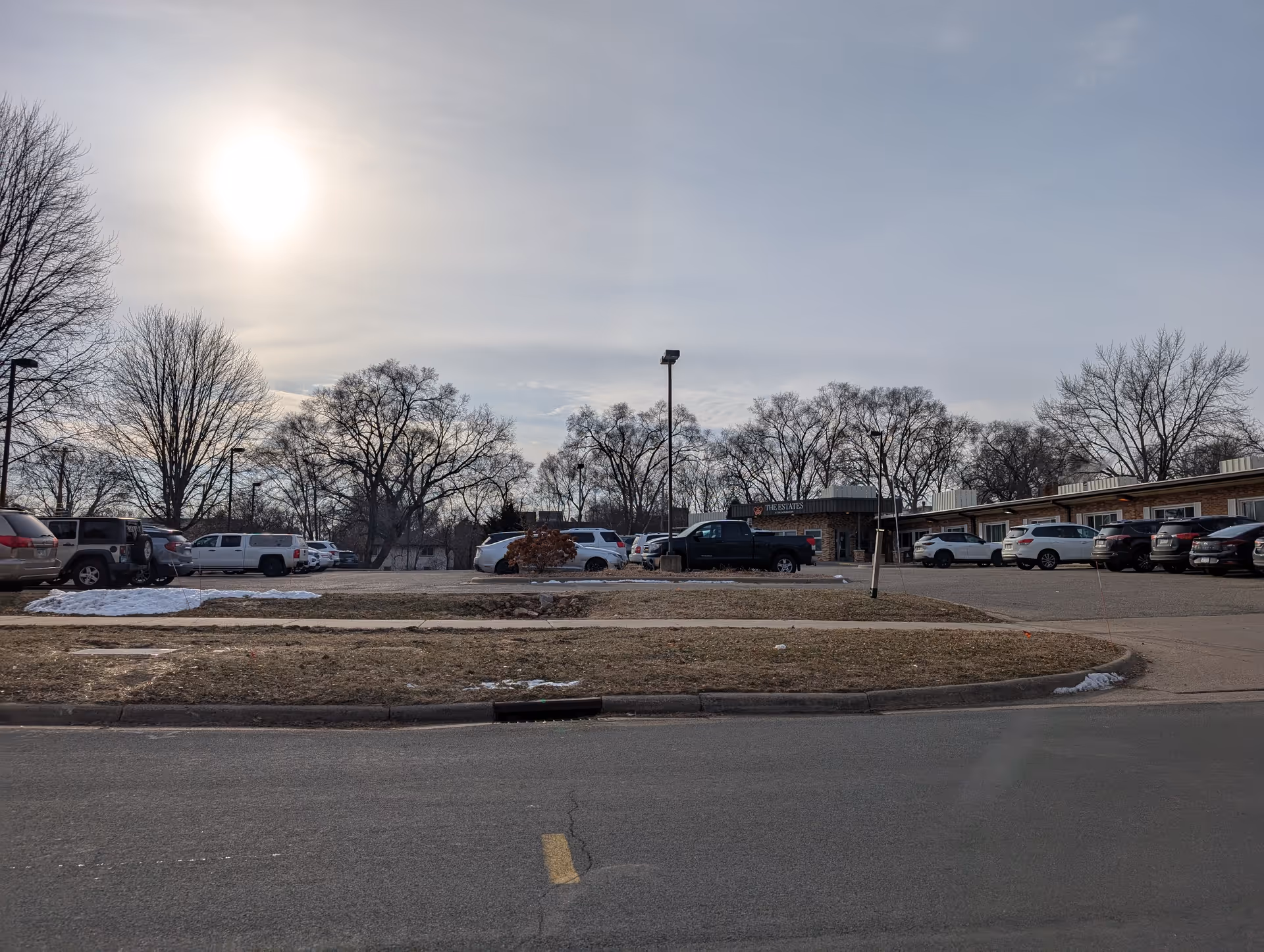 Exterior view of The Estates at Bloomington facility parking lot with several parked cars, leafless trees, and a partly cloudy sky with the sun shining.